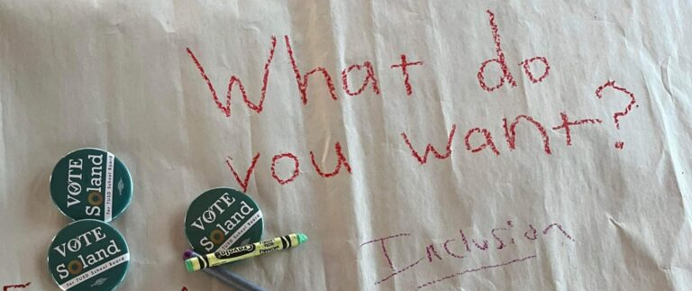 A page with the words "What do you want?" written in crayon; campaign buttons on top.